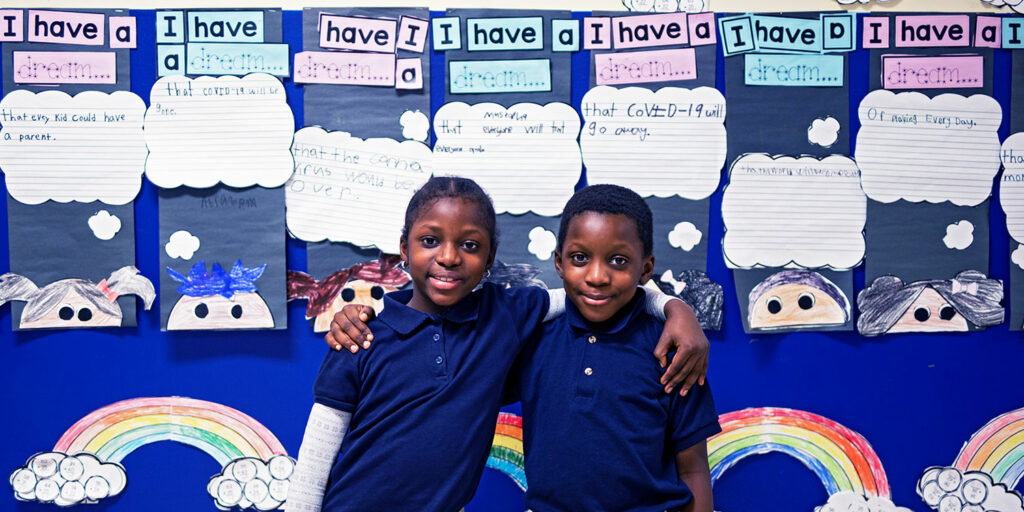 Elementary students smiling in front of bulletin board.